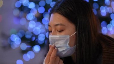 Covid-19 yüz maskesi takmış, kameraya bakan üzgün, hasta Asyalı genç bir kadının portresi. Depresyondaki kadın Noel arifesinde evin arka planında poz veriyor.