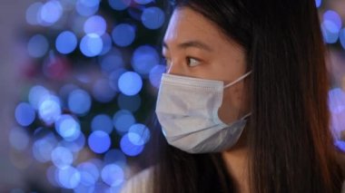 Koronavirüs maskeli hasta Asyalı genç kadın yüzünü çevirmiş üzgün bir yüz ifadesiyle kameraya bakıyor. Yılbaşı arifesinde arka planda poz veren mutsuz kadın.