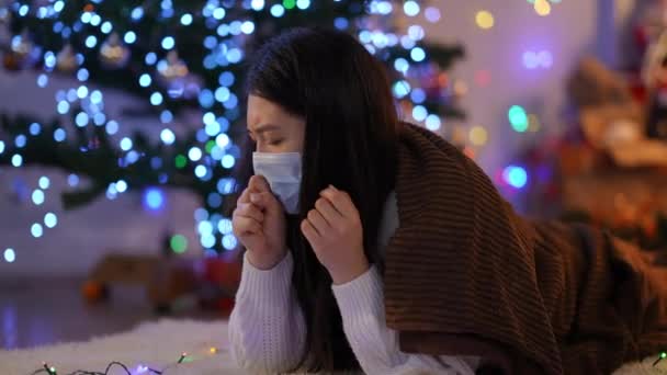 Covidフェイスマスクの悲しい病気のアジアの女性は 自宅で大晦日の温度計で体温を測定咳をします コロナウイルス感染症の症状を持つクリスマスの動揺の病気の女性 — ストック動画