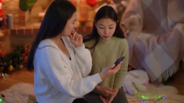 Akıllı telefon uygulamasında internette sörf yapan genç ve çekici Asyalı kadınlar yılbaşı arifesinde evde oturup gülerek konuşuyorlar. Mutlu iki arkadaş Noel kutlamasının tadını çıkarıyor.