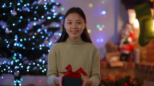亚洲纤细迷人女人伸展礼品盒的正面肖像 看着坐在圣诞灯下的相机 新年快乐 有牙齿支撑的女士 — 图库视频影像