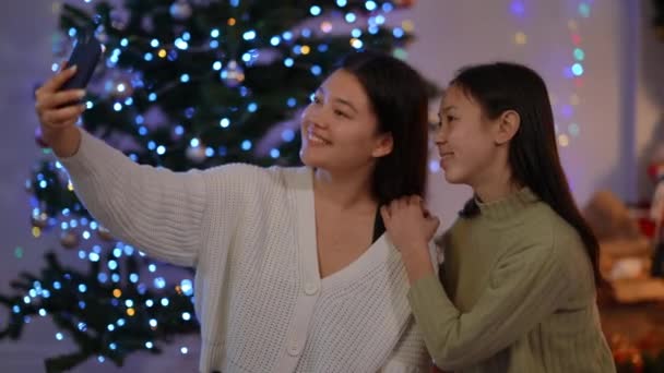 若い幸せなアジアの女性は屋内クリスマス前夜に自画自慰を振ってキスをジェスチャー空気をつかむ スマートフォンアプリで撮影する魅力的な正の女性の友人は大晦日に笑みを浮かべて — ストック動画