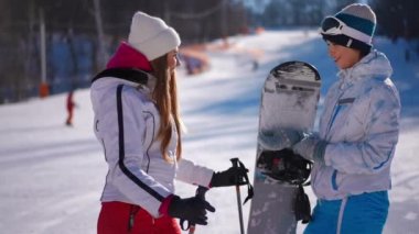 Kışın kayak ve snowboard yaparken gülen iki beyaz kadının yan görünüm portresi. İyi gevşemiş arkadaşlar, tatilin tadını çıkartın, dedikodu yapın.