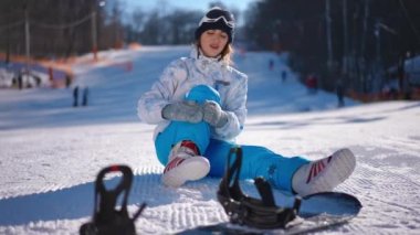 Geniş açılı, yaralı bacağını okşayan beyaz kar üzerinde oturan ve önünde snowboard yapan genç bir kadın. Güneşli bir günde, dizleri ve ayakları ağrıyan güzel beyaz bir bayanın ön görüntüsü.