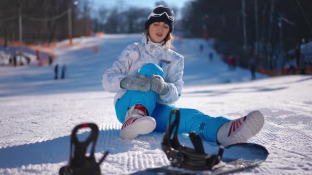 白い雪の上で前のスノーボードで負傷した足を愛撫ワイドショット若い女性 晴れた日にはスキー場で痛みを伴う膝や足を持つ美しい白人女性のフロントビュー — ストック動画