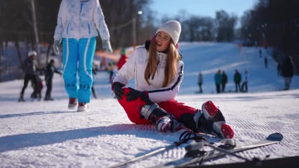 冬のリゾート地の白い雪の上に座っている負傷した膝を持つ若い女性の肖像画 秋の後に立ち上がる友人を助ける白人スリム女性 — ストック動画
