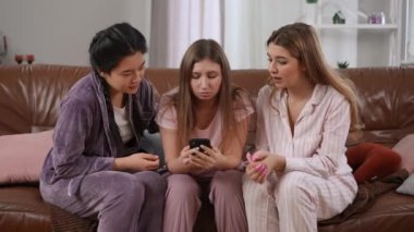 Asyalı ve beyaz kadınlar pijama partisinde kanepede oturan üzgün arkadaşına destek oluyor. Akıllı telefon mesajı bekleyen genç ve güzel arkadaşların portresi.