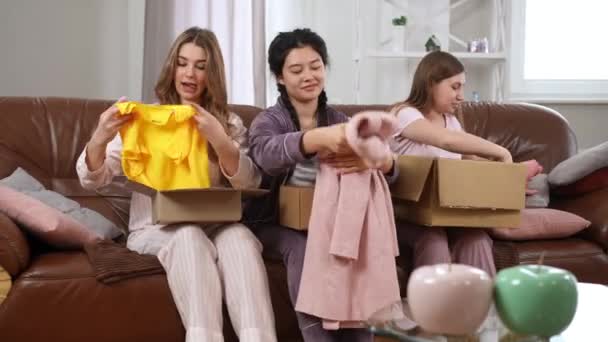 肯定的な若いアジア人と白人の女性は パジャマでソファに座って箱に古い服を詰めます 美しい女性の友人が屋内で話して寄付を集める チャリティーとライフスタイル — ストック動画