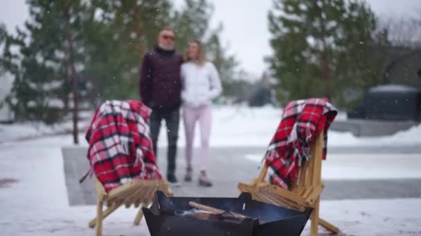 特写镜头篝火 白雪飘落 一对成年夫妇在木制椅子的背景下走来走去 白种人男子和女子在寒冷的冬日外出散步 — 图库视频影像