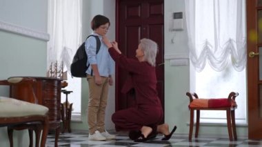 Geniş açılı, olgun bir anne, oğullarının tişörtlerini düzeltiyor, çocuklara sarılıyor ve kapıyı açıyor. Gülümseyen beyaz kadın ve çocuk sabah okula hazırlanıyor.