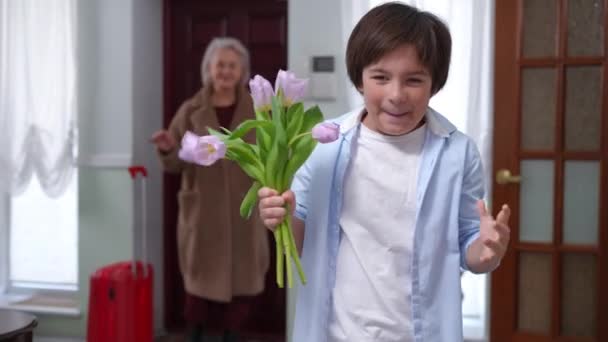 白种人迷人的儿子高兴地站在右边 捧着一束鲜花 回头望着迷蒙的母亲回家 一个笑容满面的男孩看着室内相机的画像 — 图库视频影像