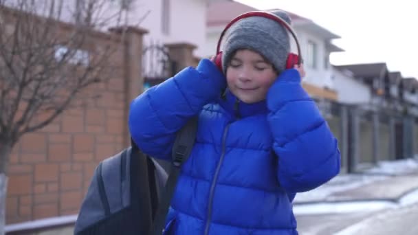多莉戴着耳机在街上边走边听音乐的无忧无虑的快乐学童的肖像 冬天放学回家 白种人可爱的男孩尽情享受着这种业余爱好 — 图库视频影像