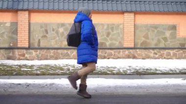 Kış günü okuldan eve koşan neşeli bir çocuğun yan görüntüsü. Geniş açılı mutlu, kendinden emin, beyaz, okul çantalı öğrencinin portresi okuldan sonra dönüyor.