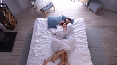 Geniş açılı mutlu çift yatakta uzanıp öpüşerek konuşuyor. Kafkasyalı kadın ve erkek rahatça yatak odasında keyif çatıyor.