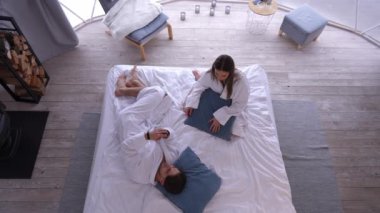 Top view, akıllı telefon uygulamasında oynayan genç bir adamı yatağa uzanmış yastık fırlatan bir kadın olarak özümsedi. Geniş açı, beyaz tenli oyuncu koca ve rahatsız bir eş yatak odasında.