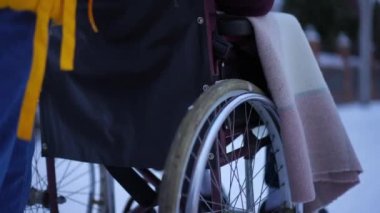 Dışarıdaki karlı yolda ağır çekimde oturan bir adamla tekerlekli sandalye iten tanınmayan bir kadın. Hasta adamla ilgilenen kendine güvenen genç bir bayan kış akşamı şehir caddesinde geziniyor.