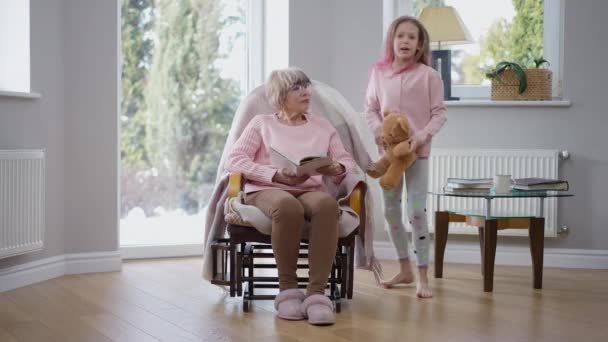 Радісна Безтурботна Мила Дівчина Бігає Старшій Жінці Читаючи Книжку Вітальні — стокове відео