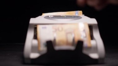 Erkek parmak banknot sayacının başlama düğmesine basıyor ve yakın plan hesaplanan Euro banknotlarını bozuyor. Siyah arkaplanda paralı bir aygıt