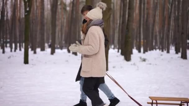 在冬季森林谈话中 正面女孩和男孩拉着雪橇散步的侧视图跟踪镜头 广博自信的白人朋友在公园里享受户外休闲 童年与友谊 — 图库视频影像