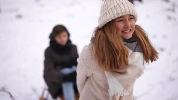 一个年轻的白人女孩拉着雪橇和男孩坐在冬季森林里的肖像 可爱迷人的少年试骑雪橇在公园度假的朋友 — 图库视频影像