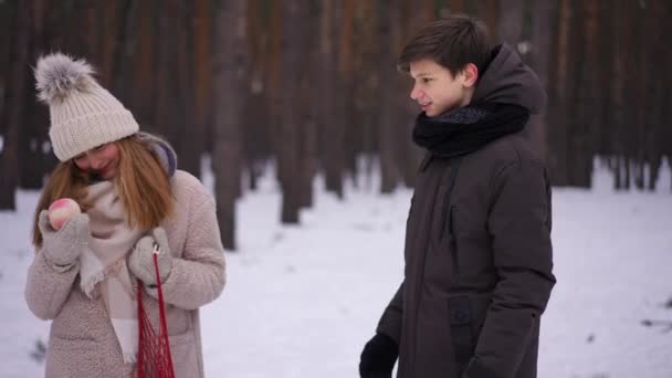 かわいい10代の女の子は冬の森の中で立って話して男の子においしい桃を渡す 10代の白人カップルの肖像画日付休暇で公園で 第一の愛と関係概念 — ストック動画