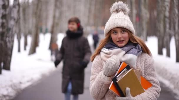 背景にはぼやけた少年をバックに放課後本を片手に歩く聡明な10代の少女 スマート白人のティーンエイジャーの肖像 友人と一緒に冬の公園を散歩 — ストック動画