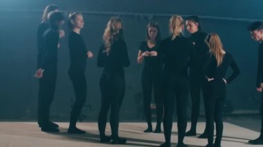 Siyahlı beyaz erkek ve kadınların sahnede durup gülümseyerek konuşmalarının geniş açılı portresi. Mutlu kendinden emin oyuncular ve oyuncular içeride tiyatro performansı prova ediyorlar. Eğleniyorlar.