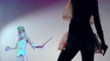 Beyaz elbiseli, ince, güzel bir kadın. Önünde duran, kılıçlı, siluet gibi bir kadın. Sahnede sahne ışıkları altında sahne alan beyaz aktrisin portresi.