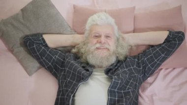 Gülümseyen, beyaz saçlı, uzun saçlı, sakallı ve ağır çekimde poz veren adamın portresini büyüt. Mutlu kaygısız Kafkasyalı erkek emekli evde yatakta uzanmış kameraya bakıyor.
