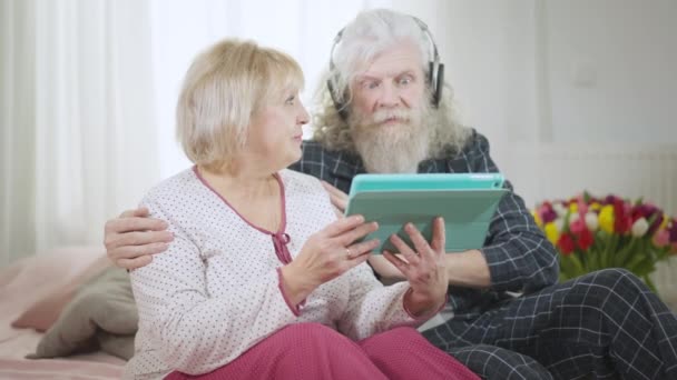 满意的白种人老年夫妇穿着睡衣坐在家里 在网上听音乐 自信的微笑着的男人和女人一起享受周末在屋里聊天的乐趣 慢动作 — 图库视频影像