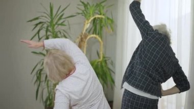 Back view senior Caucasian man and woman in pajamas exercising in the morning in bedroom. Motivated confident couple training at home indoors in slow motion