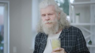 Yakışıklı, beyaz, sağlıklı vitamin içeceği içen, kameraya bakarak el hareketi yapan bir adam. Kendine güvenen emeklinin portresi. Lezzetli içkinin tadını çıkarıyor. Sabahları evde ağır çekimde poz veriyor.