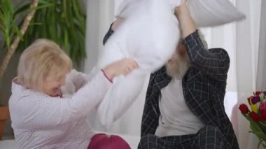 Evdeki yatakta oturup yastık kavgası yapan kaygısız iki emekli. Kahkahalarla gülen beyaz bir erkek ve kadın yatak odasında eğleniyor. Yaşam tarzı ve eğlence konsepti