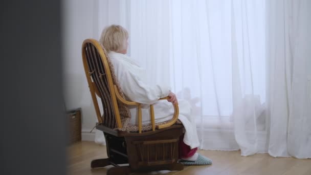 身穿白色浴衣的白人妇女坐在摇椅上向窗外看去 放松无忧无虑的女性退休人员在家里客厅里享受着早晨的生活 慢动作 — 图库视频影像