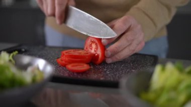Close-up senior male hands cutting red tomato for vegetable salad with knife. Unrecognizable Caucasian male retiree cooking tasty healthful breakfast at home in kitchen