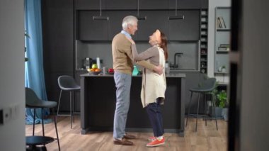 Wide shot loving smiling senior husband dancing with wife spinning spouse indoors. Happy confident Caucasian couple of retirees enjoying leisure at home in kitchen
