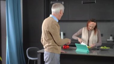 Positive confident senior man talking with woman cooking tasty healthful salad and turning looking at camera smiling. Portrait of happy Caucasian husband enjoying morning with wife at home