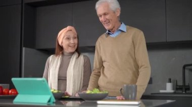 Couple of senior bloggers waving greeting subscribers in social media using digital tablet app. Positive smiling Caucasian man and woman cooking in kitchen talking streaming online