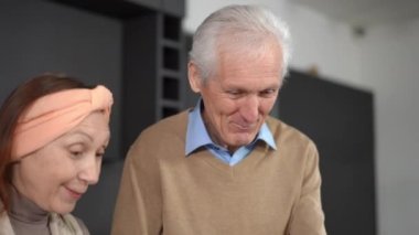 Portrait of smiling handsome senior husband feeding joyful wife with cucumber slice in kitchen at home. Happy loving Caucasian couple cooking breakfast having fun in the morning