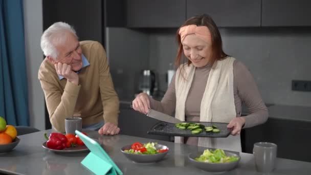 快乐的老年女性烹调蔬菜沙拉 与男人和丈夫交谈 赞美可爱的妻子 快乐自信的白种人夫妇退休后在厨房享用早餐 — 图库视频影像