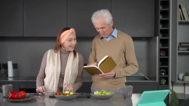 家庭でおいしい健康的なサラダを調理するレシピを読んで幸せな高齢者のカップル 陽気な白人男性と女性の中出し肖像室内で食材を準備話して — ストック動画