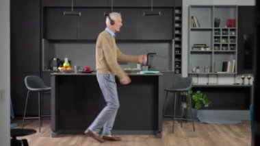 Wide shot positive senior man dancing moonwalk having fun in kitchen at home. Happy carefree Caucasian male retiree in headphones listening to music enjoying leisure indoors