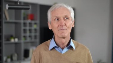 Panning portrait of grey-haired confident senior man with grey eyes thinking looking away drinking morning coffee from cup. Handsome Caucasian male retiree enjoying morning at home in kitchen
