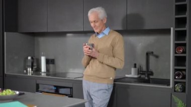 Portrait of thoughtful Caucasian senior man drinking coffee in the morning standing in kitchen at home. Handsome male retiree enjoying hot drink indoors looking away dreaming
