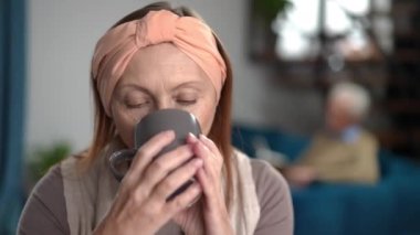 Close-up portrait of charming happy senior woman enjoying tasty morning coffee with blurred man sitting at background. Relaxed carefree Caucasian female retiree drinking hot drink smiling