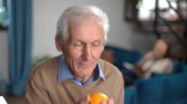 Smiling senior man smelling vitamin orange looking at camera. Portrait of positive happy Caucasian male retiree posing in kitchen indoors with blurred wife at background