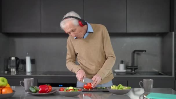 快乐的资深白人男子在家里的厨房里用耳机跳舞烹调沙拉 快乐无忧无虑的退休人士在室内准备蔬菜配料的肖像 — 图库视频影像