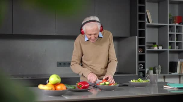 积极乐观的白种人老年人切蔬菜沙拉跳舞的音乐 快乐的无忧无虑的退休人员 带着耳机 在家里的厨房里享受着乐趣 — 图库视频影像