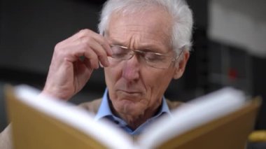 Close-up front view of concentrated senior man reading book taking off putting on eyeglasses turning page. Focused Caucasian interested male retiree enjoying literature at home indoors