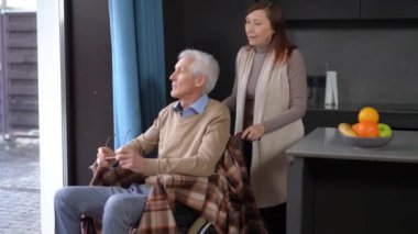 Senior Caucasian woman pushing wheelchair with man to window talking smiling. Loving caring wife enjoying leisure with husband at home indoors. Bonding and lifestyle